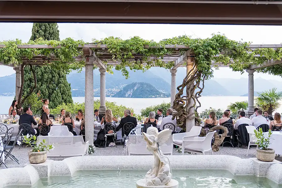 Outdoor wedding dinner on Lake Como under a pergola with fountain in the foreground and lake view.