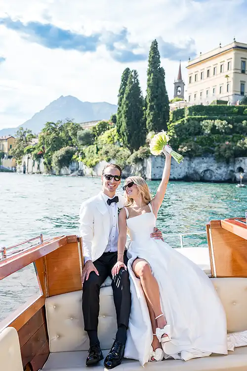 Bride and groom on a wooden boat during a luxury wedding on Lake Como with villa gardens in the background.