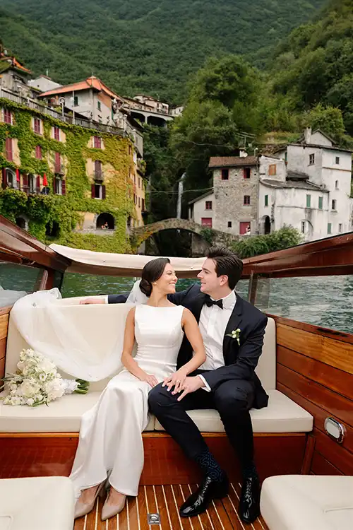 Bride and groom on a wooden boat on Lake Como with a historic lakeside village in the background.