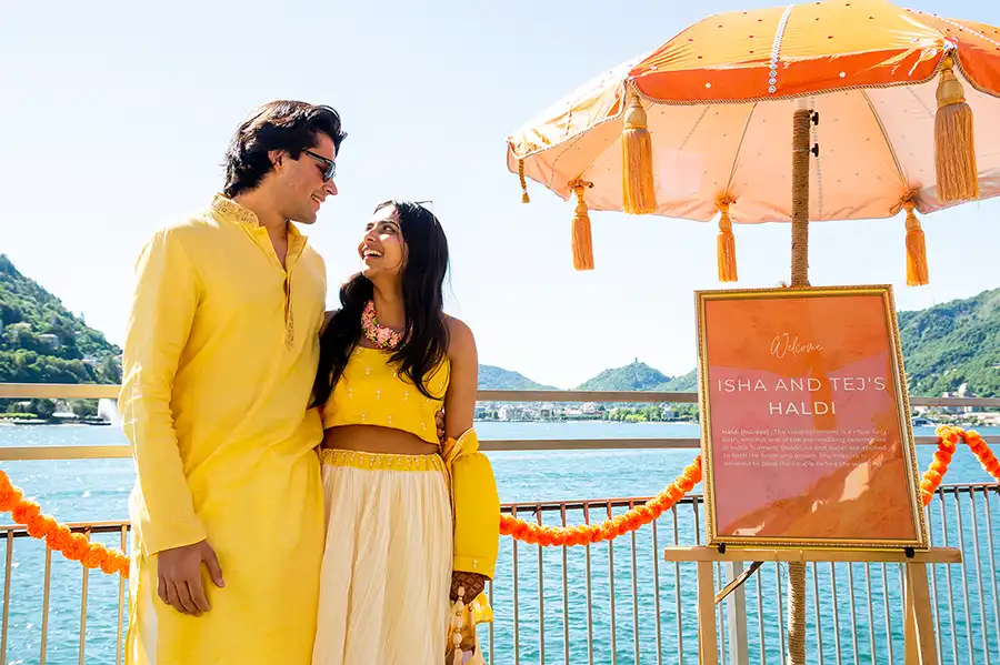 Indian pre-wedding celebration on Lake Como with the couple in yellow outfits beside a Haldi welcome sign.