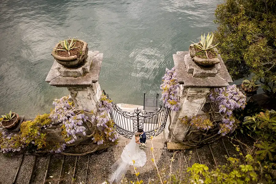 Bride and groom photographed by the lake on Lake Como beside a historic gate framed by wisteria.
