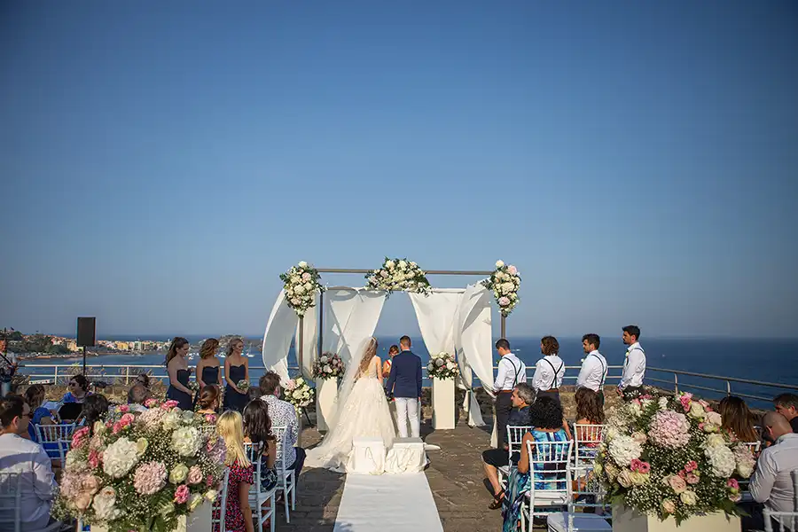 Seaside wedding ceremony in Sicily with white aisle, floral arrangements and panoramic coastal view