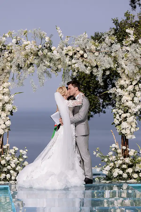 Bride and groom kissing under a white floral wedding arch with sea view in Sicily